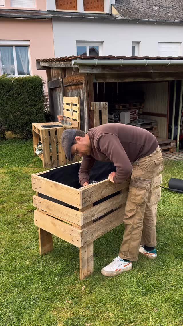 Man assembling a large square planter box made from wooden pallet slats outdoors.