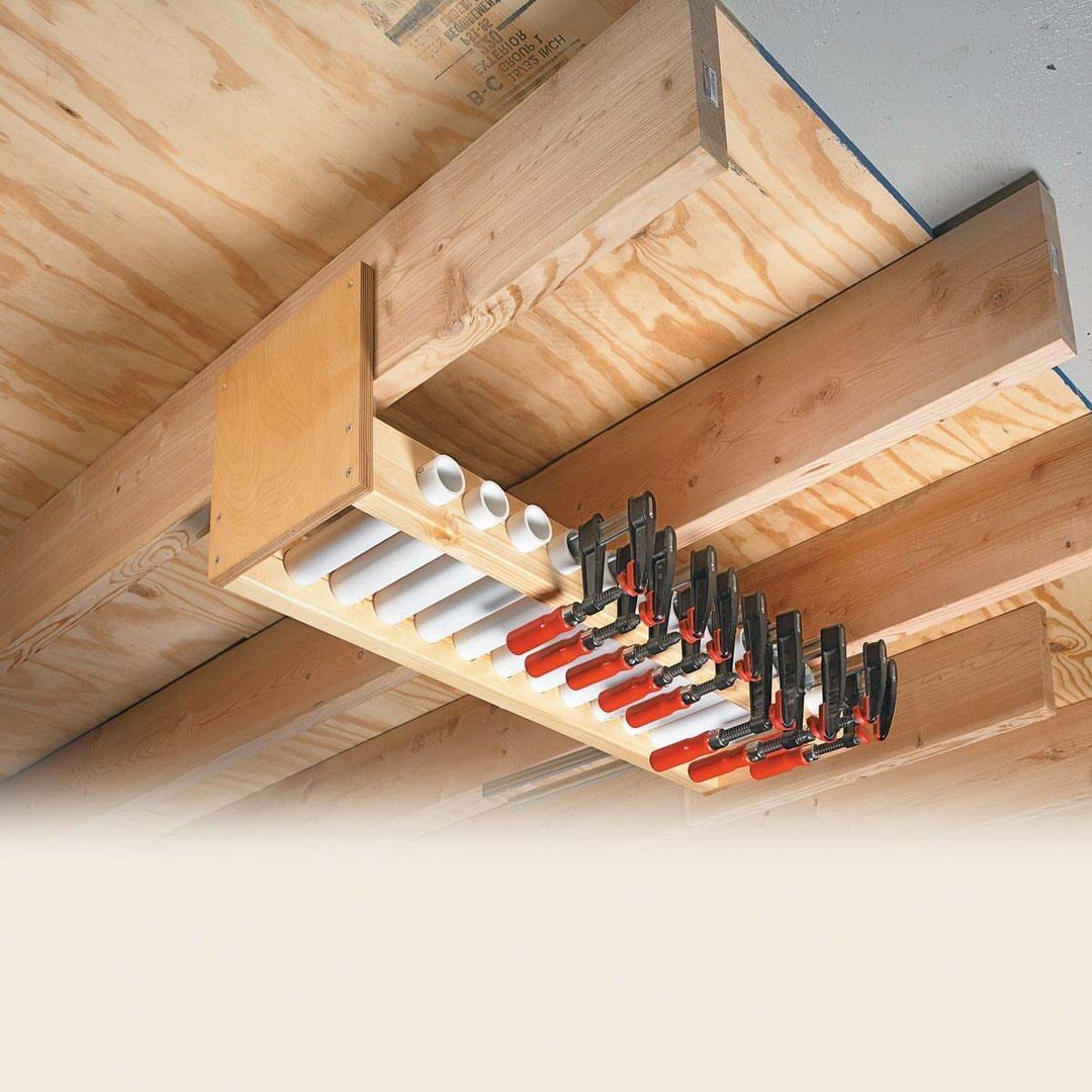 8 Woodshop Design Ideas for Smarter Storage and Layout 1 Wooden ceiling rack holding red and black bar clamps and white PVC pipes.