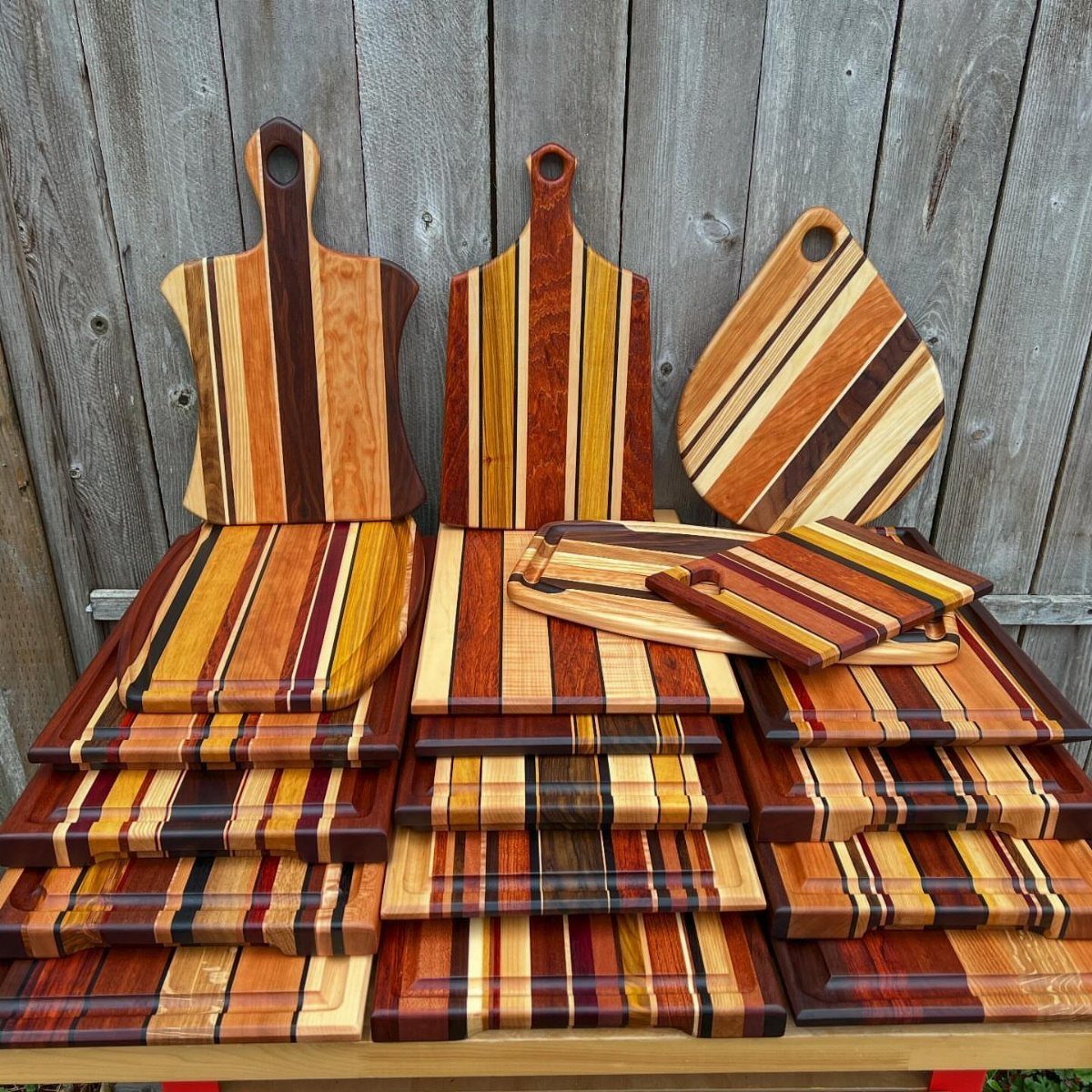 Assorted striped wooden cutting boards of various shapes leaning against a grey wooden fence.