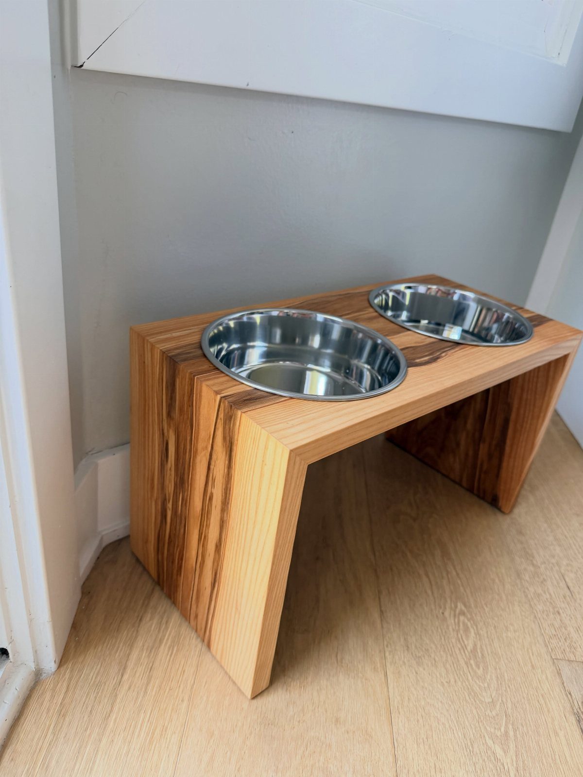 Wooden dog bowl stand with two stainless steel bowls on a light floor.