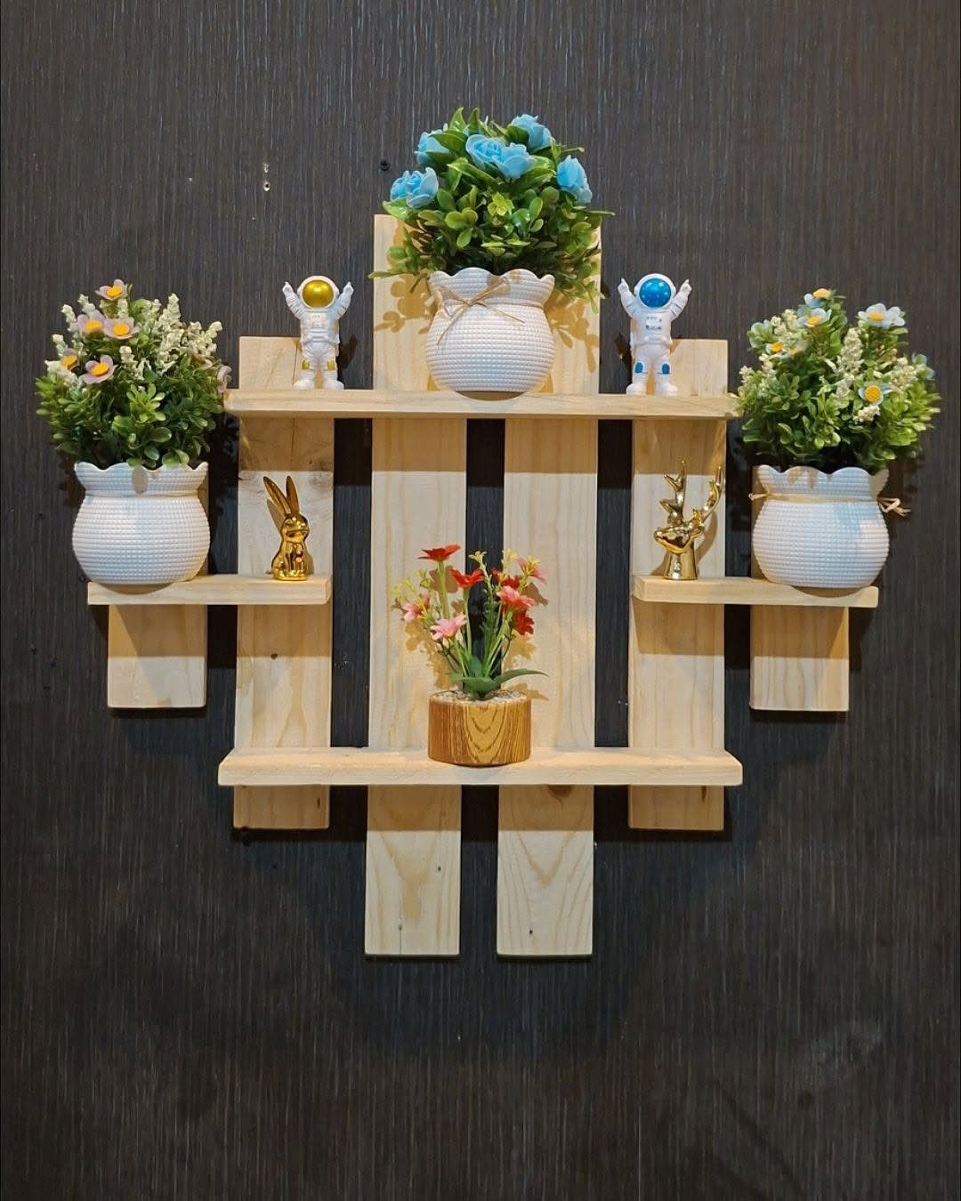 Wooden wall shelf unit holding small potted plants and decorative figurines against dark wall.