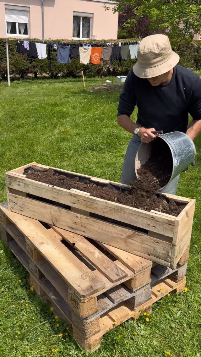 Man filling a pallet wood planter box with soil in a green yard.