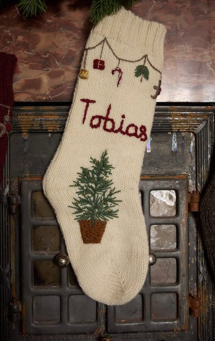 Cream knitted Christmas stocking with name Tobias and tree hanging on a fireplace
