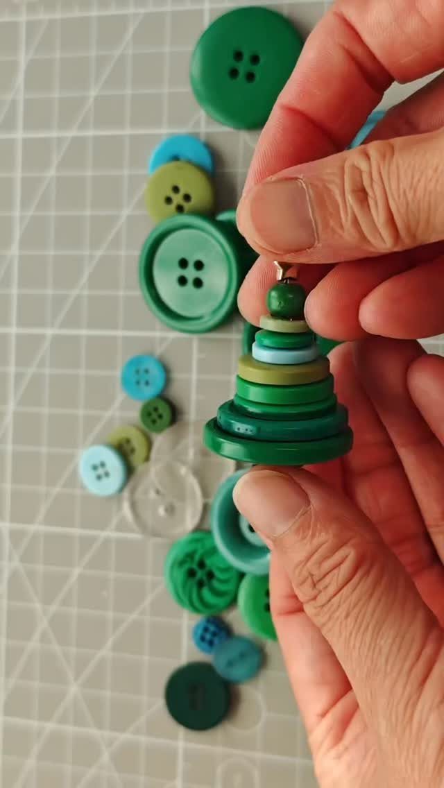 Hands assembling a small Christmas tree ornament made from stacked green and blue buttons.